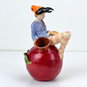 CARRIANNE L HENDRICKSON woman with beak hat sitting on apple sculpture 2…